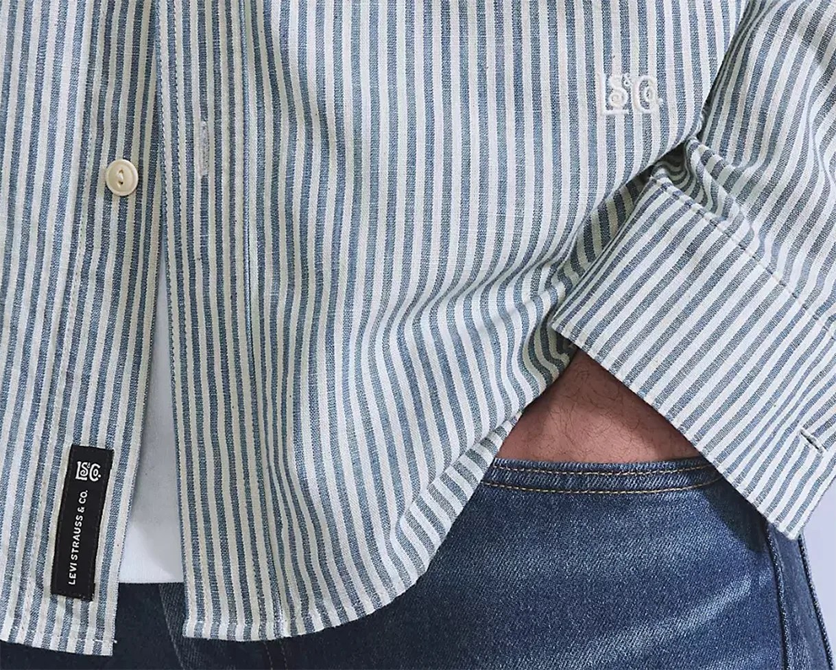 Close-up of a person wearing a vertically striped button-up shirt in light blue and white, partially tucked into blue denim jeans. The shirt has a small embroidered "LS&Co." logo on the chest and a black fabric tag near the bottom hem that reads "LEVI STRAUSS & CO." The person's hand is tucked into the front pocket of the jeans.