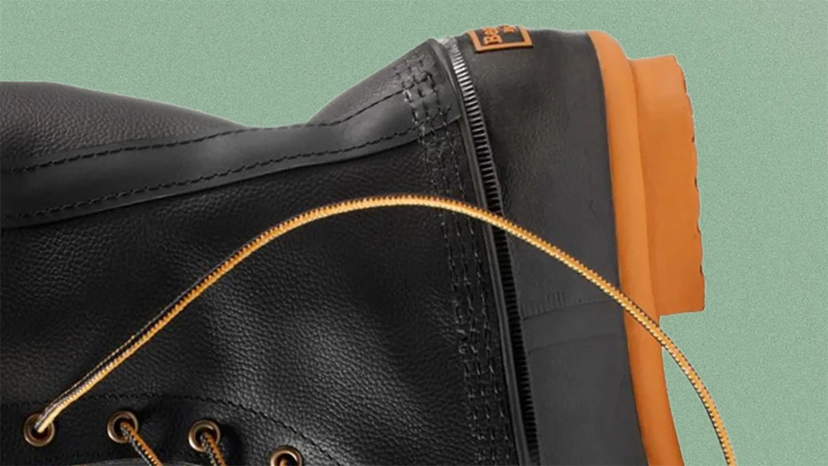 Close-up of a black leather boot with a textured surface, featuring yellow-brown laces threaded through metal eyelets. The boot has a thick, orange rubber sole with a rugged tread pattern. The stitching and construction details are visible, highlighting the boot's durability.