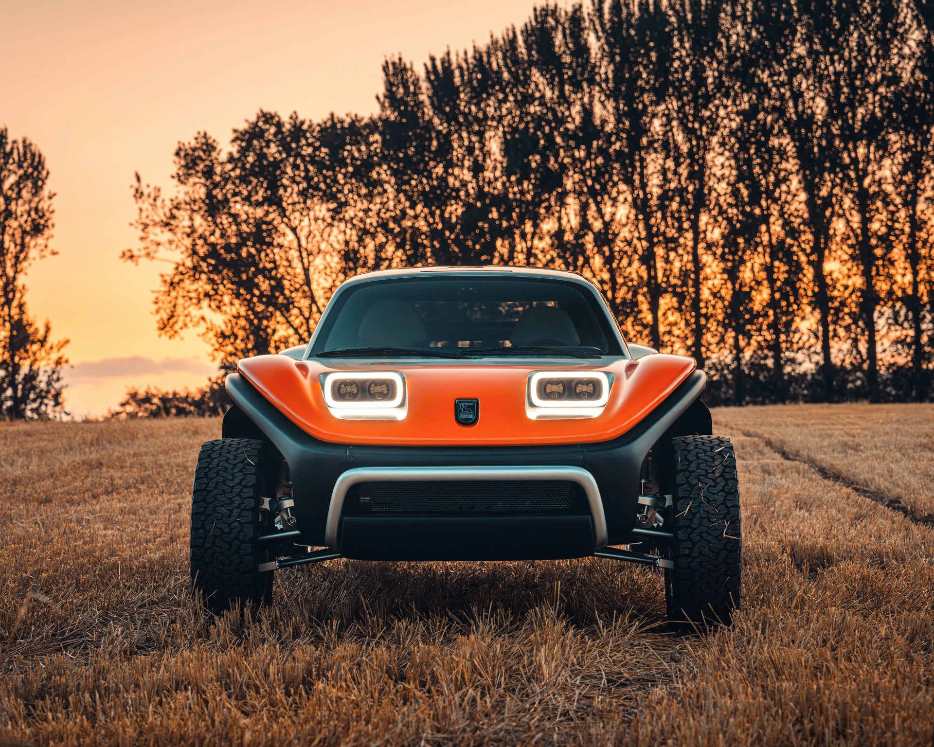 Front view of an orange and black off-road vehicle with large tires, illuminated square headlights, and a rugged design, parked on a dry grassy field at sunset with tall trees in the background.