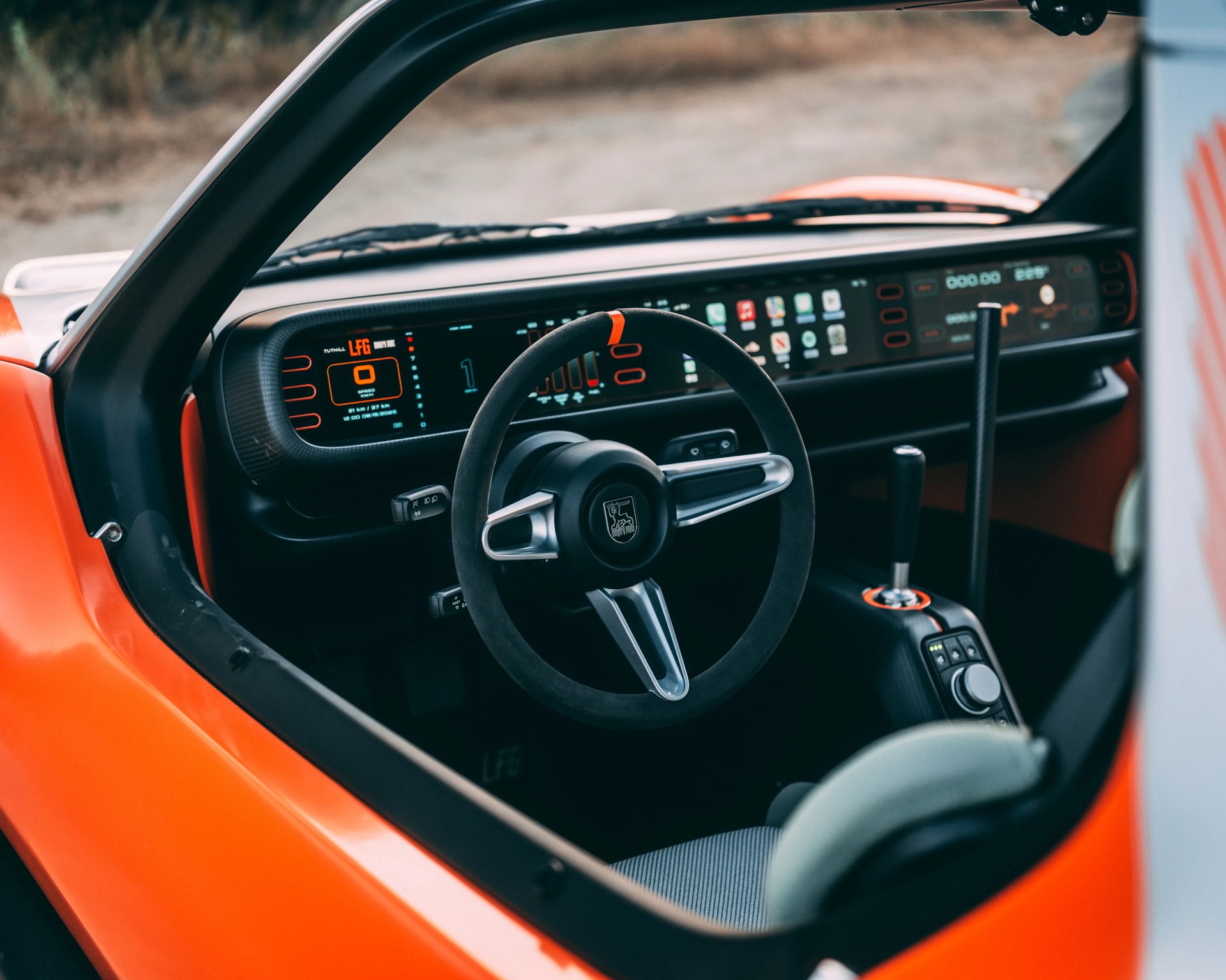 Interior view of a modern car featuring a black steering wheel with silver accents and an emblem in the center. The dashboard has a wide digital display panel with orange and blue graphics, showing speed, gear, and various icons. The car has an orange exterior visible around the door frame, and the center console includes a gear lever and control buttons. The seat is partially visible with a gray fabric cover.