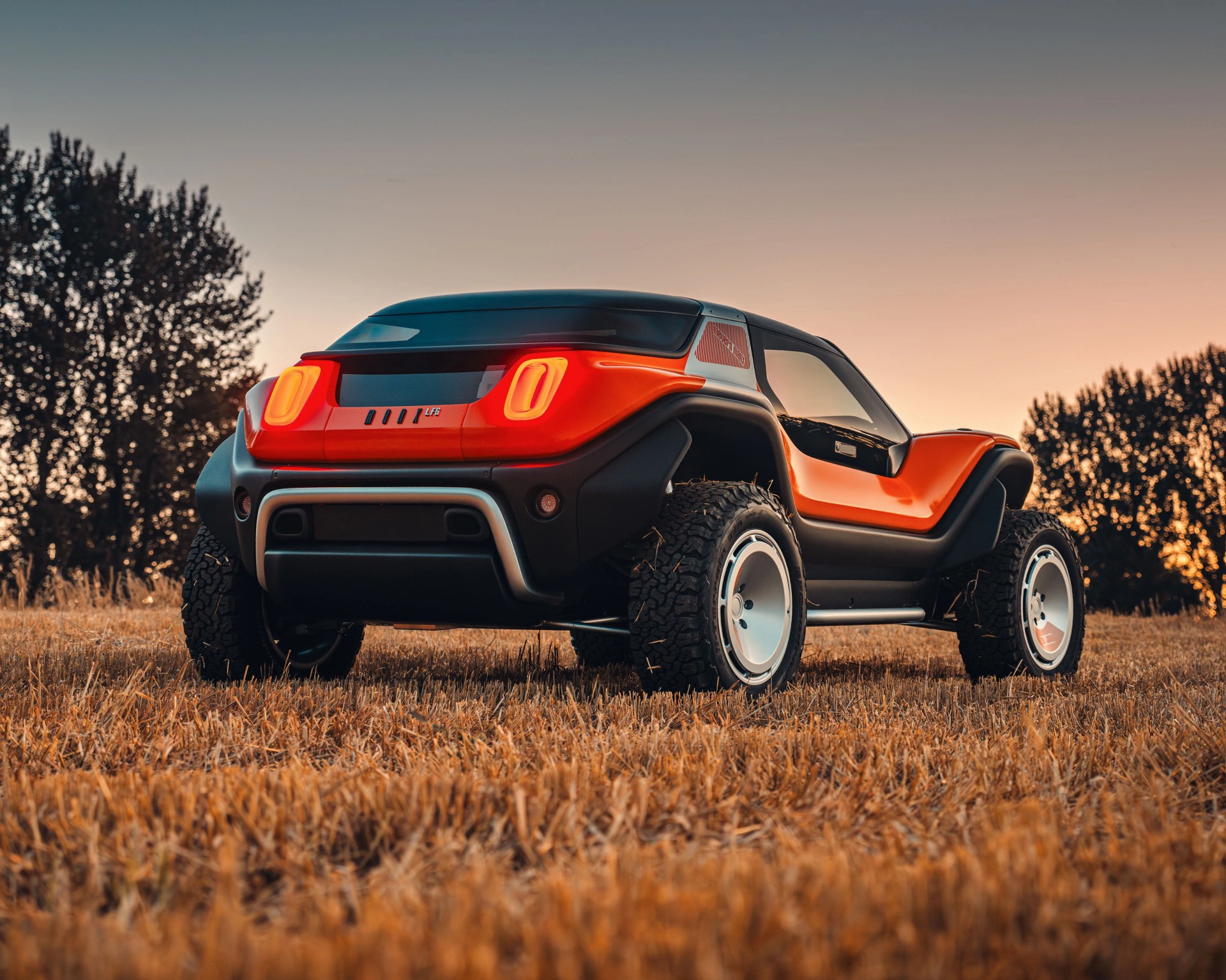 A compact, futuristic off-road vehicle with a bright orange and black body, large rugged tires, and distinctive rear lights, parked on a dry grassy field at sunset with trees in the background.
