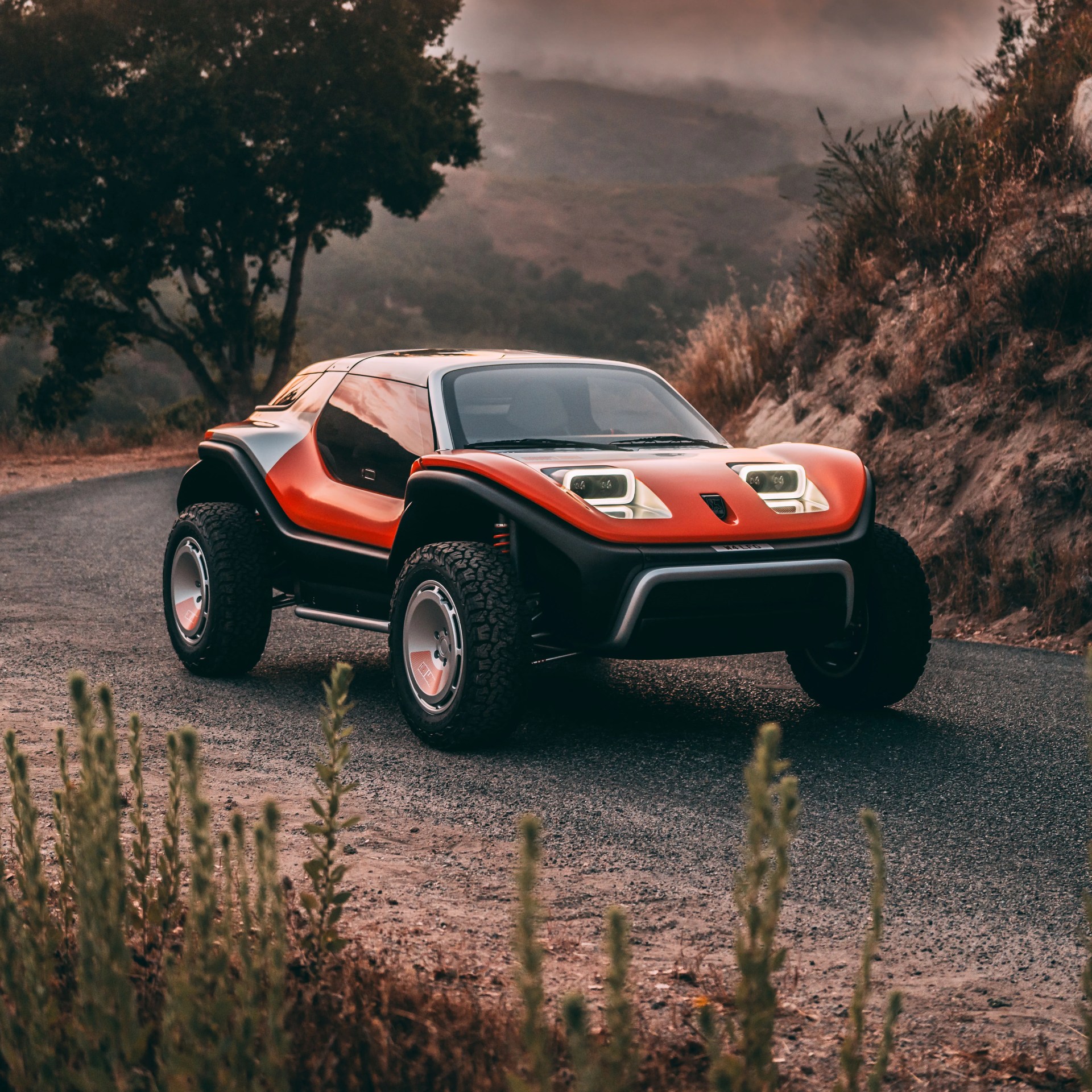 A compact, futuristic off-road vehicle with a sleek, aerodynamic design. It features a bright orange and black body, large rugged tires, and distinctive rectangular headlights with LED accents. The car is parked on a winding road surrounded by natural scenery, including trees and dry grass.