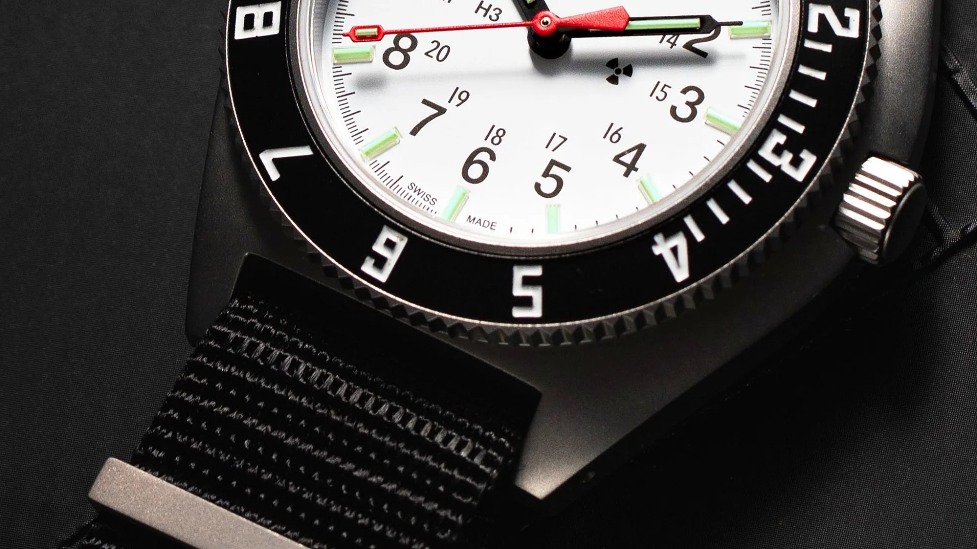 Close-up of a wristwatch with a white dial, black bezel with white numerals, and a black fabric strap. The watch hands are red and green, with luminous green hour markers. The dial includes black Arabic numerals for hours and smaller numbers for 13 to 20, along with the text "SWISS MADE" near the 6 o'clock position. The bezel has a textured edge, and the crown is silver with ridges.