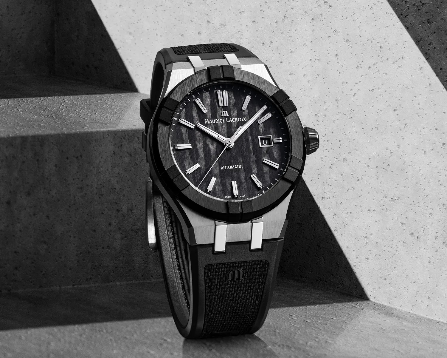 Maurice Lacroix automatic wristwatch with a black textured dial, silver hour markers, and a date window at 3 o'clock. The watch has a black bezel with a brushed finish and a black strap featuring a textured pattern and the Maurice Lacroix logo. The background is a concrete surface with strong diagonal shadows.