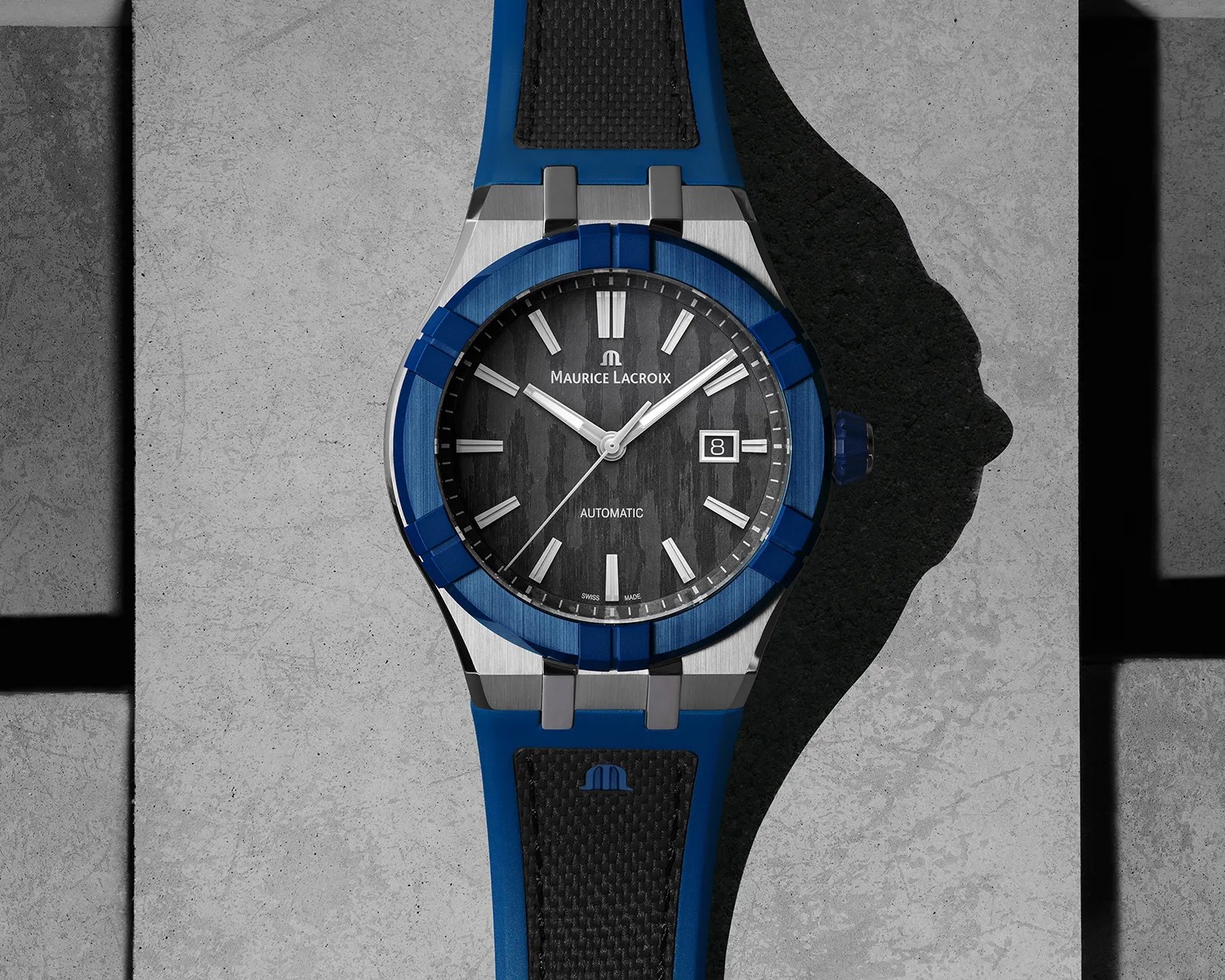 A Maurice Lacroix wristwatch with a blue bezel and blue rubber strap featuring a black textured center. The watch face is black with silver hour markers and hands, including a date window at the 3 o'clock position. The strap has a black textured insert with a blue Maurice Lacroix logo. The watch is placed on a gray concrete surface.