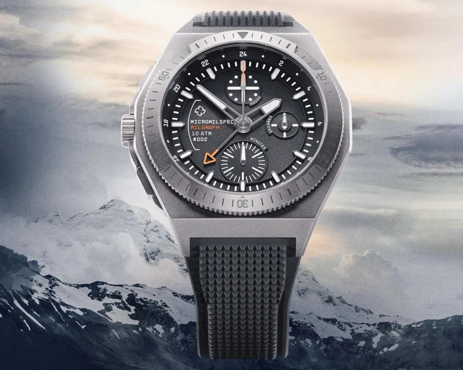A rugged wristwatch with a black textured dial featuring white hour markers and hands, and an orange arrow-tipped hand. The watch has three subdials labeled "hours," "minutes," and "seconds." The bezel is metallic with minute markings, and the watch case is angular and metallic gray. The strap is black with a textured pattern. Text on the dial reads "MICROMILSPEC MILGRAPH 10 ATM #002." The background shows a mountainous landscape under a cloudy sky.