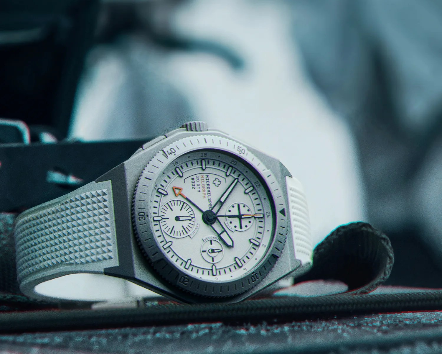 A close-up of a wristwatch with a white textured rubber strap and a matte gray case. The watch face is white with black markings and features three subdials, bold black hands, and an orange arrow-tipped hand. The bezel has minute markers, and the dial includes text reading "MICROmilspec MILgraph 20 ATM #002." The watch is resting on a textured surface with a blurred background.