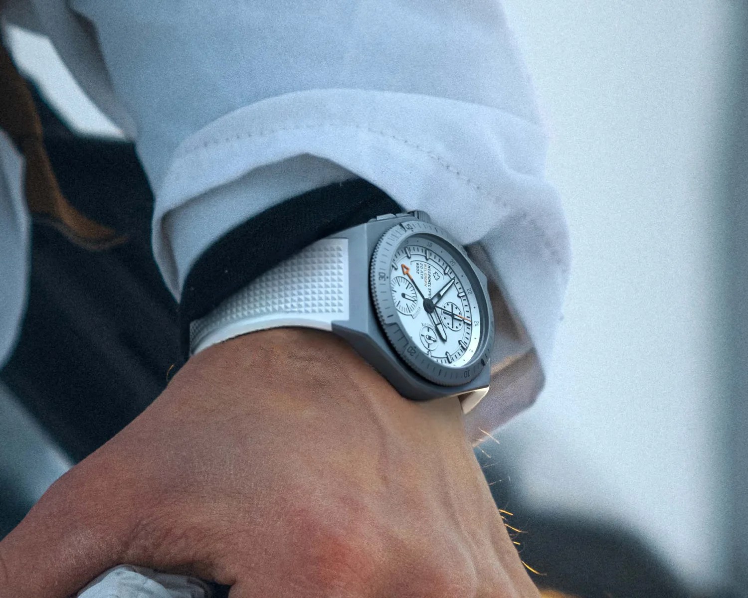 Close-up of a micromilspec wristwatch with a white textured rubber strap and a silver bezel, worn on a wrist with a light blue shirt cuff. The watch face is white with black and orange details, featuring multiple subdials and a date window.