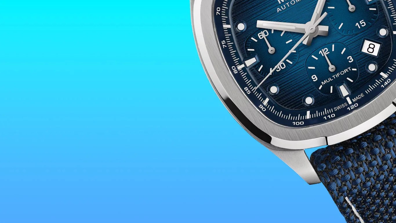Close-up of a luxury mido wristwatch with a blue textured dial featuring three subdials, white hour markers, and a date window at the 3 o'clock position. The watch has a polished silver bezel and a blue woven fabric strap. The dial includes the text "MULTIFORT" and "SWISS MADE.