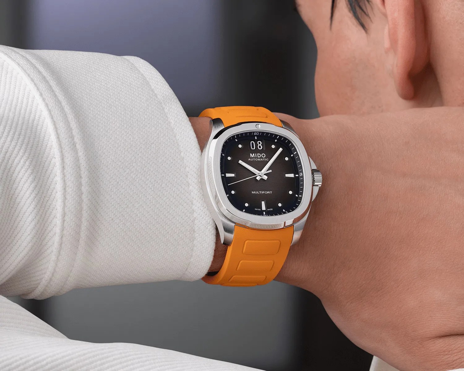 A wristwatch with a square stainless steel case and a black dial featuring white hour markers and hands. The watch has a date display at the 12 o'clock position and the words "MIDO AUTOMATIC" and "MULTIFORT" on the dial. It is worn on a wrist with an orange rubber strap. The wearer is dressed in a white textured long-sleeve garment.