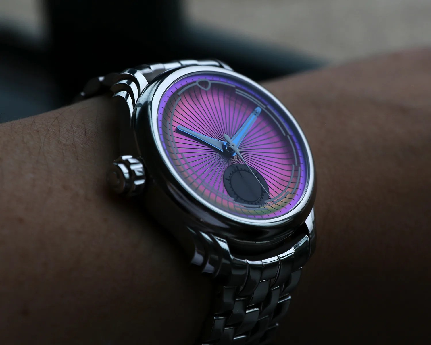 A ming wristwatch with a metallic silver band and case, featuring a vibrant purple dial with radiating lines. The watch has blue hour and minute hands, a thin silver second hand, and a small black subdial near the 6 o'clock position. The watch is worn on a person's wrist.
