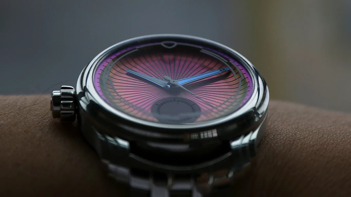 Close-up of a ming wristwatch with a metallic case and a purple-pink gradient dial featuring radiating black lines. The watch has blue hour and minute hands and a small subdial near the 6 o'clock position. The watch is worn on a wrist with a metal bracelet.