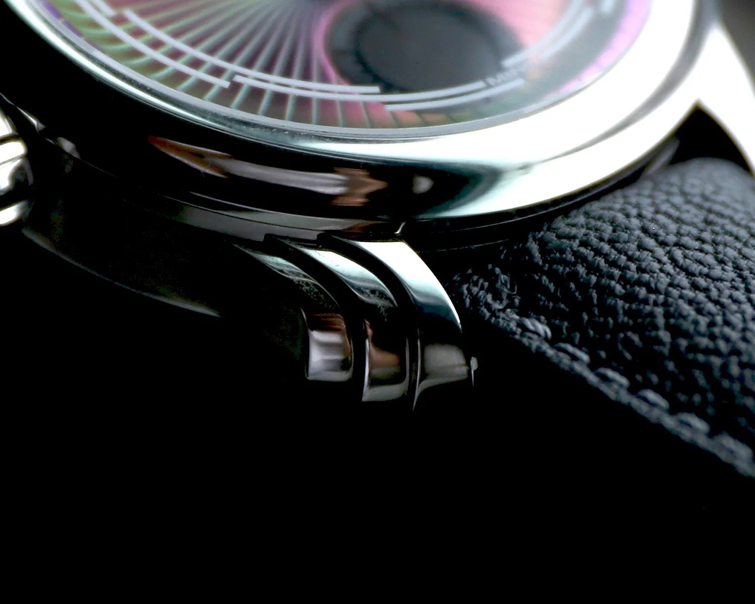 Close-up of a ming wristwatch showing the polished metal case and lugs, part of the textured black leather strap, and a portion of the watch face with fine markings and a hint of purple and pink hues.