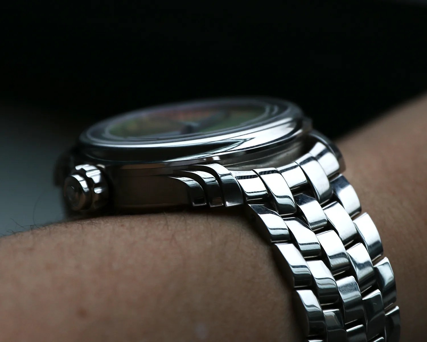Close-up of a ming wristwatch with a polished silver metal bracelet and a round case, worn on a person's wrist. The watch crown is visible on the left side of the case. The watch face is slightly blurred and not fully visible.