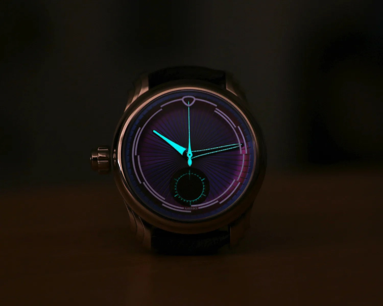 A close-up of a ming wristwatch with a glowing blue-green hour and minute hands, and a small seconds subdial at the 6 o'clock position. The watch face has a dark purple background with a radial pattern and luminous markers around the edge. The watch case is metallic with a crown on the left side.