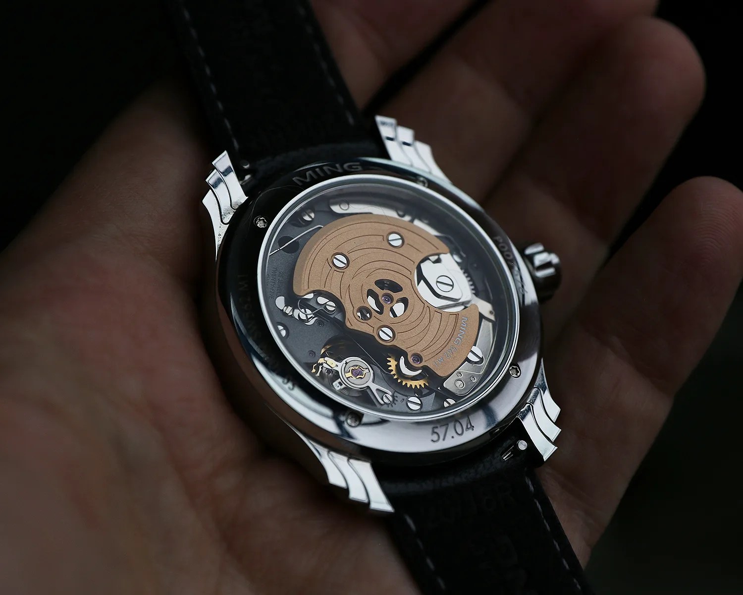 Close-up of the back of a ming wristwatch held in a hand, showcasing the intricate mechanical movement inside. The watch has a transparent case back revealing gears, screws, and a rotor with a gold-toned finish. The outer case is polished metal with the number "57.04" engraved near the bottom. The watch strap is black with visible stitching.