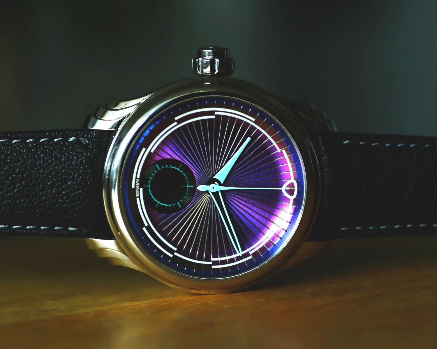A ming wristwatch with a metallic case and a black leather strap featuring white stitching. The watch face has a striking design with a purple background and radiating lines. It has light blue hour and minute hands, a small circular subdial on the left side, and white markings around the edge. The brand name "MING" is visible on the left side of the dial.