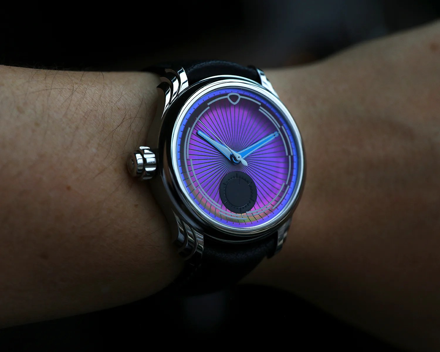 A ming wristwatch with a shiny silver case and a black leather strap, featuring a vibrant purple dial with radiating lines. The watch has blue hour and minute hands and a small black subdial near the 6 o'clock position. The watch is worn on a person's wrist.