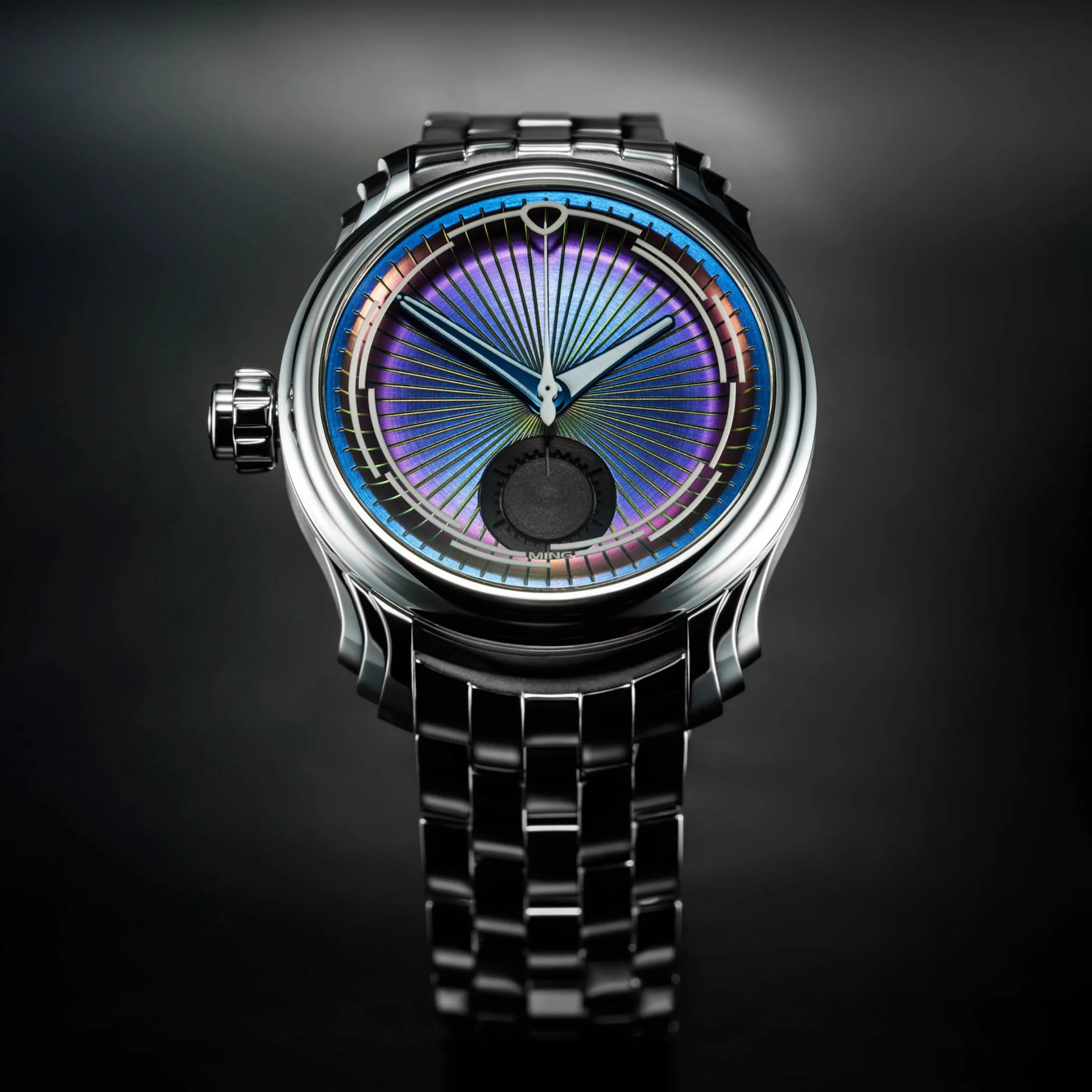 A luxury ming wristwatch with a polished silver metal bracelet and case. The watch face features a vibrant, iridescent dial with shades of purple, blue, and green, accented by fine gold radial lines emanating from the center. The hour and minute hands are silver with a blue tint, and there is a small black subdial at the 6 o'clock position. The watch crown is prominent on the left side of the case. The background is dark and blurred, highlighting the watch.
