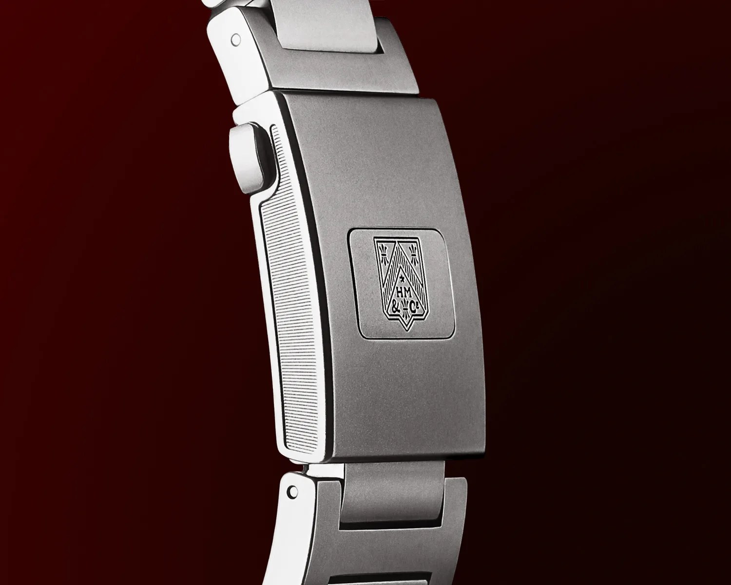 Close-up of a silver moser watch clasp with a detailed emblem featuring the letters "HM & MC" engraved on the center. The clasp has a brushed finish and a side button for release, set against a dark red background.