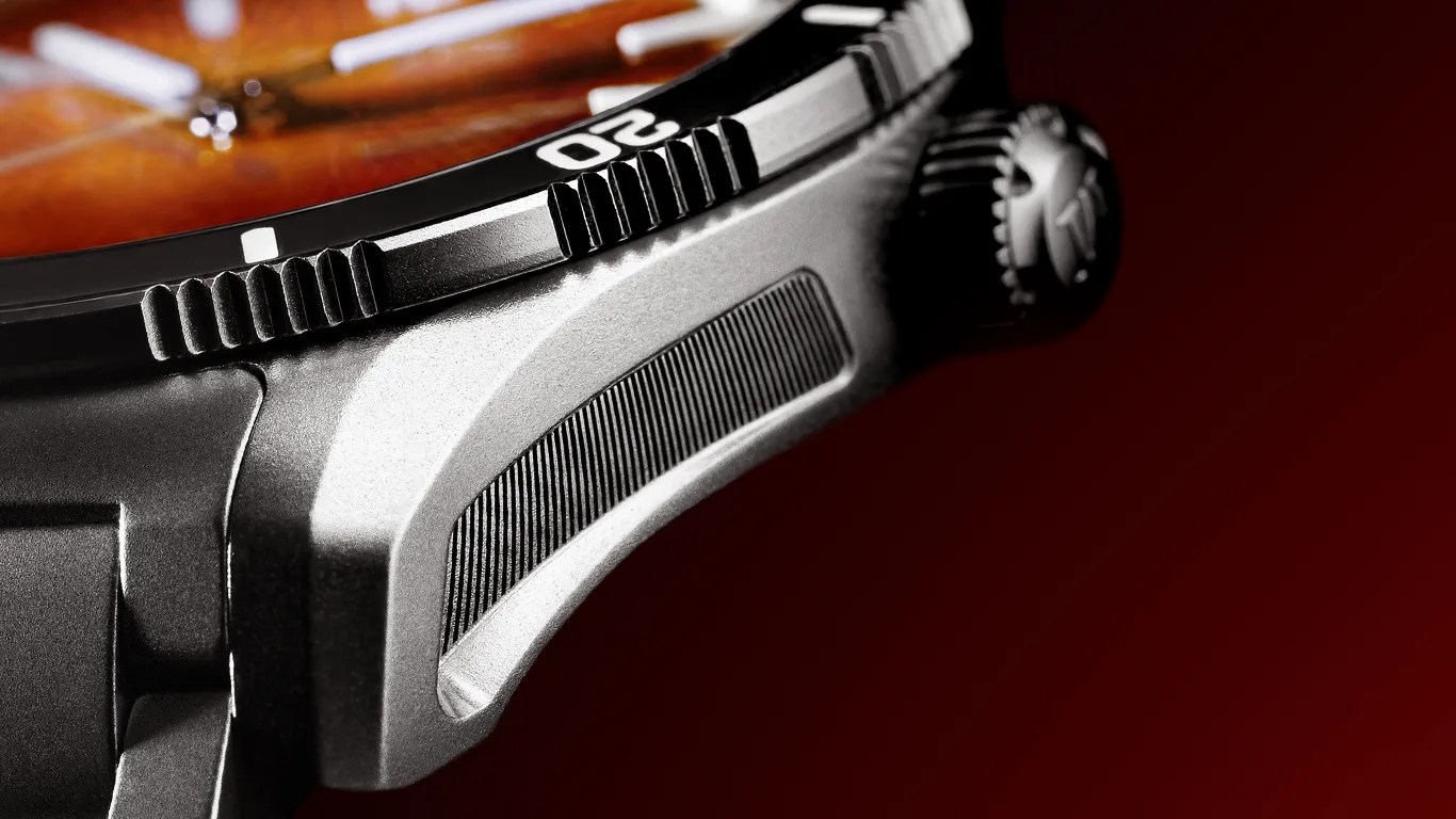 Close-up of a moser watch case showing the textured side grip, part of the bezel with the number "20," and the crown with a logo, against a dark red background. The watch face has a warm, amber tone with visible hands.