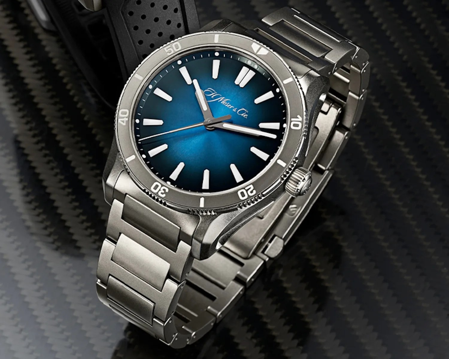 Silver metal wristwatch with a blue gradient dial, white hour markers, and a rotating bezel marked with numbers in increments of ten. The watch has a stainless steel link bracelet and is set against a carbon fiber textured background. The brand name "H. Moser & Cie" is visible on the dial.