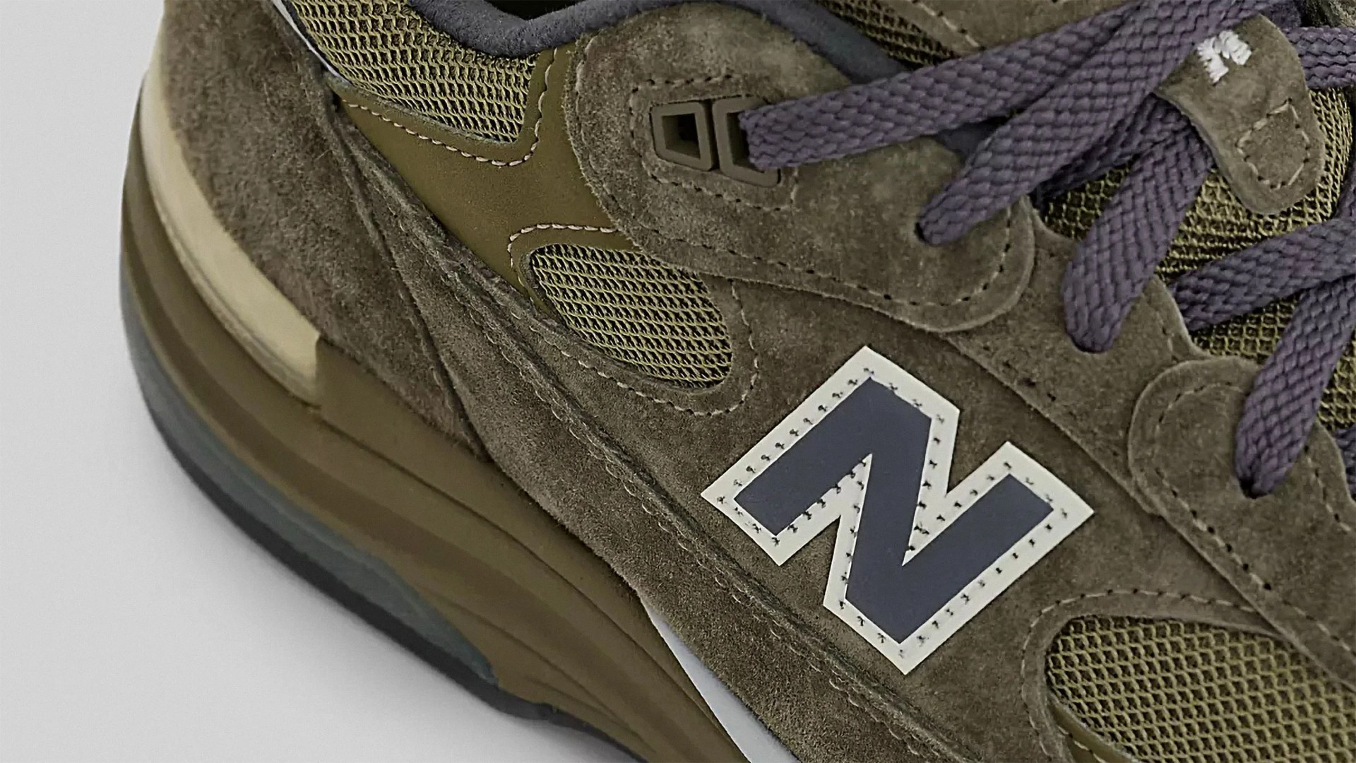 Close-up of a brown New Balance sneaker featuring suede and mesh materials, purple laces, and the signature "N" logo on the side. The sole is layered with shades of brown and beige.