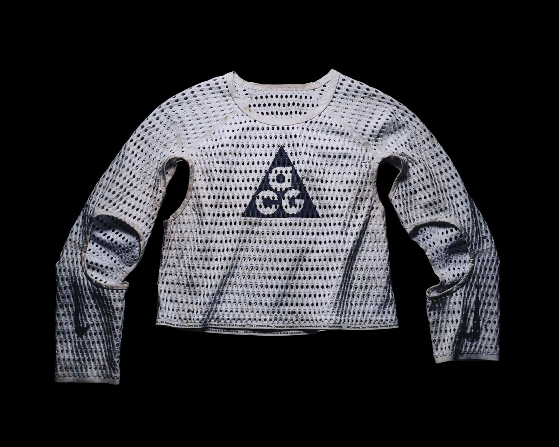 White long-sleeve mesh shirt with a pattern of evenly spaced holes throughout. It features a black triangle logo with the letters "a," "c," and "g" inside the triangle on the chest. The shirt has a round neckline and visible stitching along the sleeves and hem. The hem also has a small printed text band.