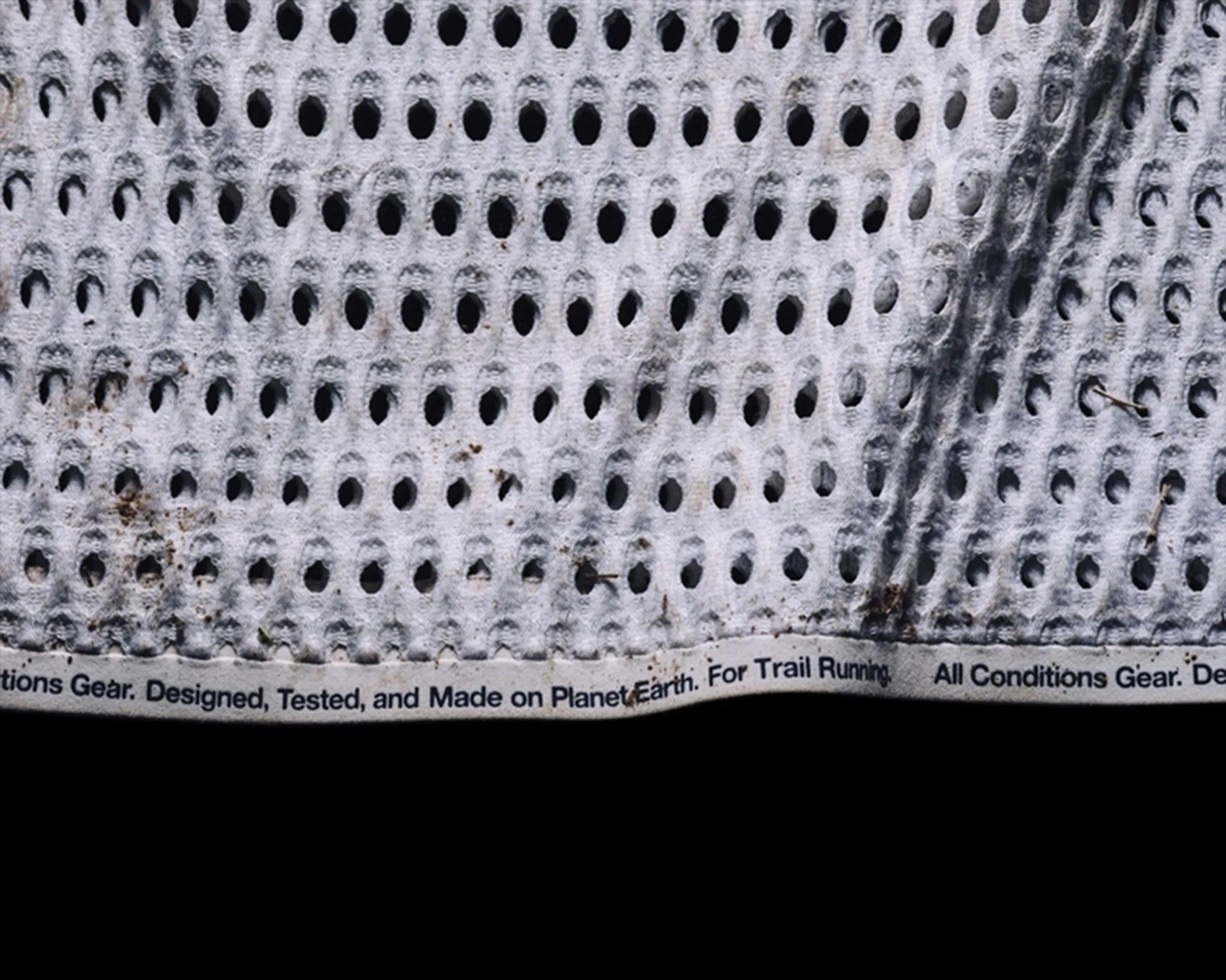 Close-up of a white, perforated fabric with dirt marks and small debris. The fabric edge has printed text that reads: "Designed, Tested, and Made on Planet Earth. For Trail Running. All Conditions Gear.
