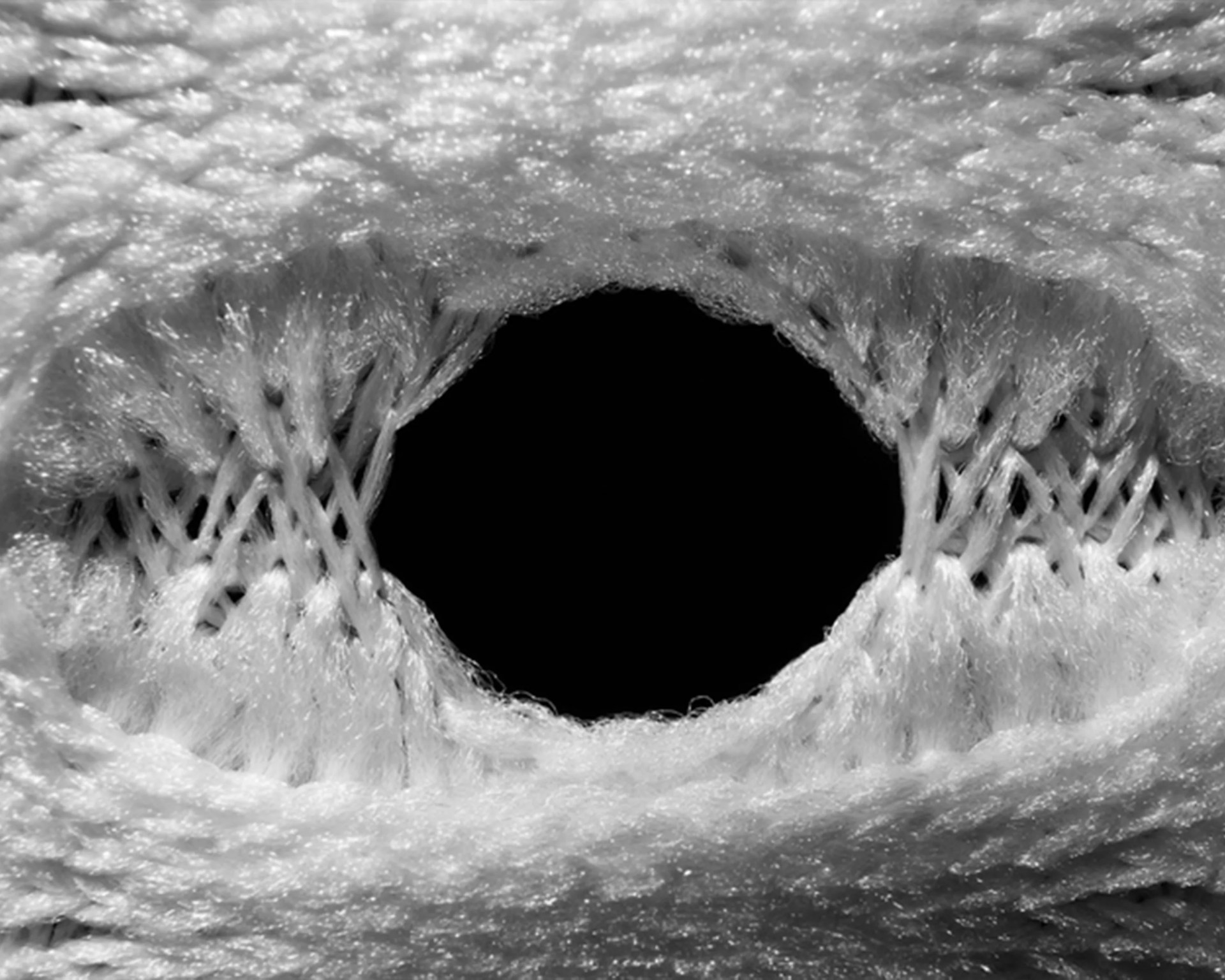 Close-up of a knitted fabric with a large, oval-shaped hole in the center, showing the individual yarn fibers and stitches around the opening. The yarn appears soft and slightly fuzzy.