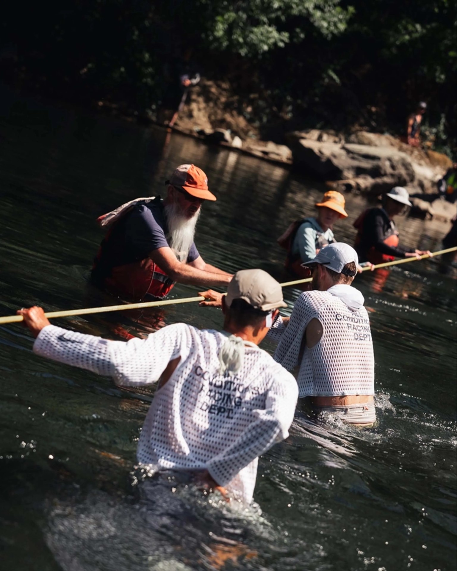 People standing waist-deep in a river, holding a long pole together. They are wearing hats and light, mesh-like shirts, with some wearing orange or red waders. The background shows rocky riverbanks and trees.