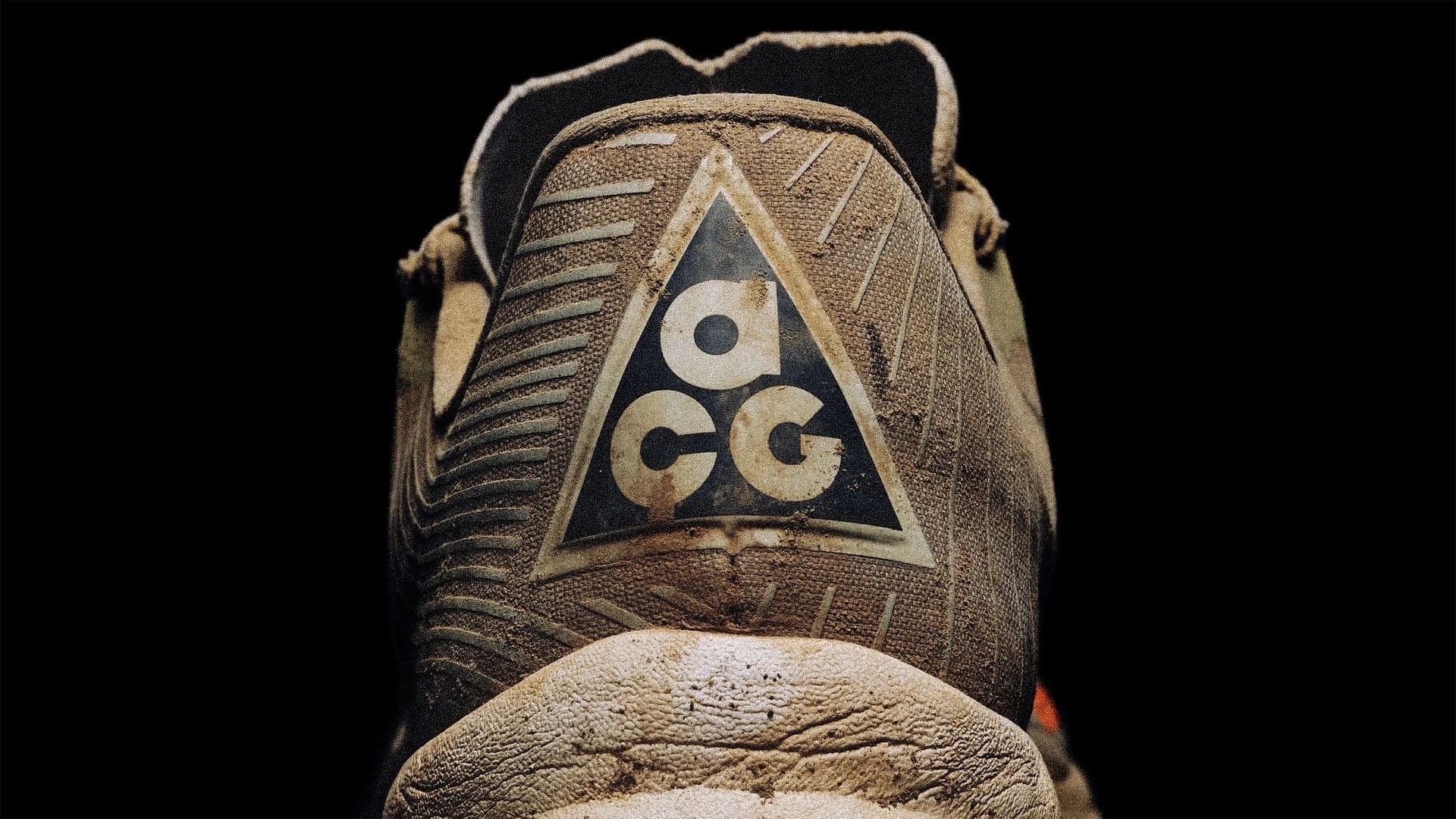 Close-up of the back heel of a worn, dirty brown sneaker with a textured surface. The heel features a triangular logo with the letters "a," "c," and "g" inside it. The background is black, highlighting the shoe.
