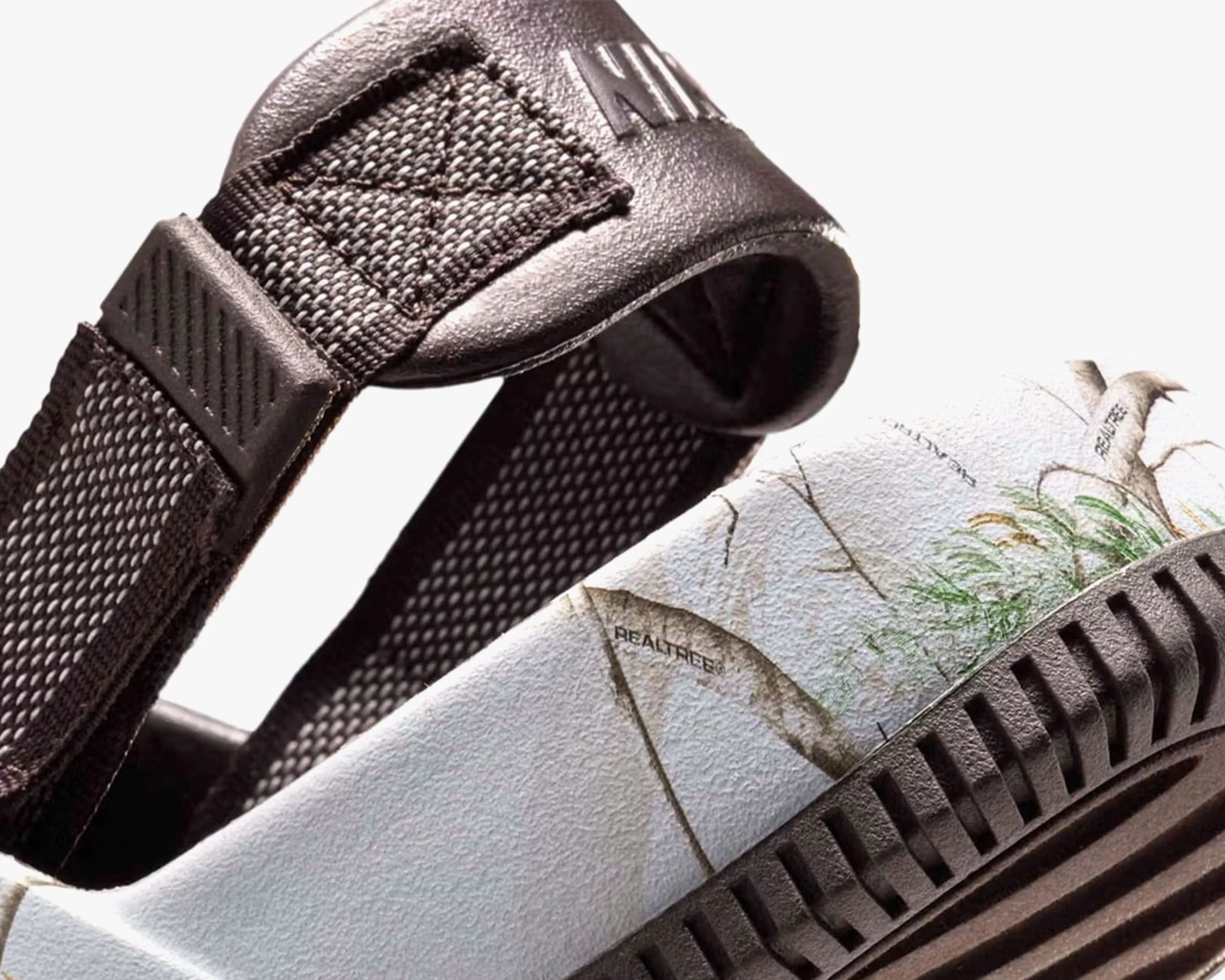 Close-up of a shoe heel featuring a camouflage pattern with grass and branches, the word "REALTREE" printed on it, and a black textured strap with reinforced stitching and a leather patch embossed with "AIR." The sole is black with a ridged design.