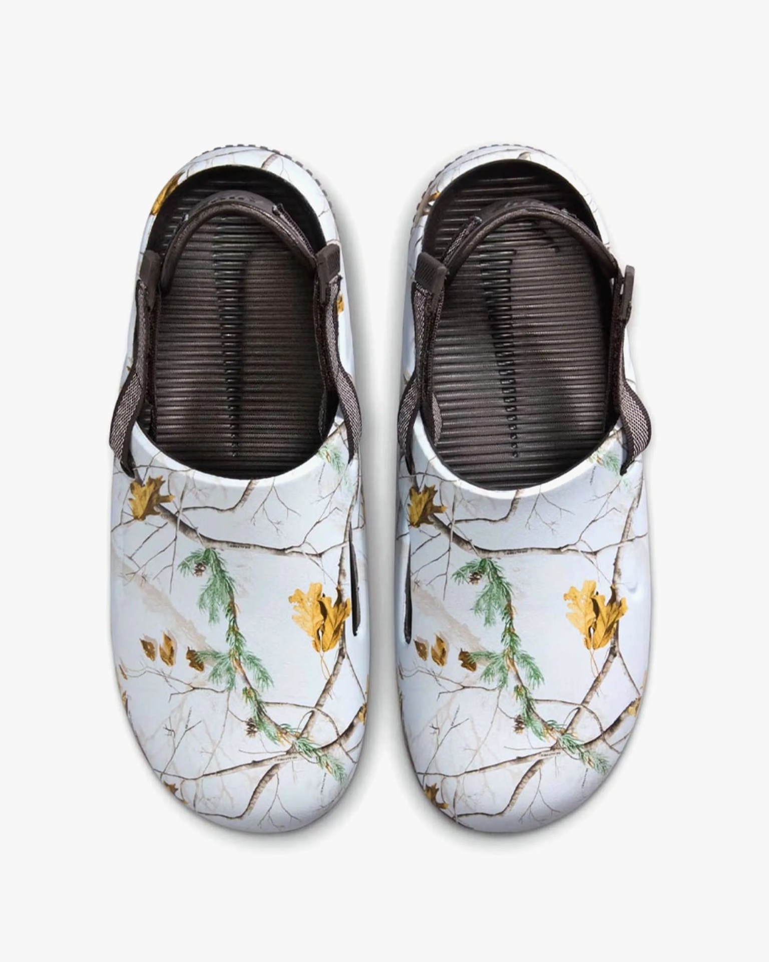 A pair of slip-on clogs with a white base featuring a nature-inspired print of brown branches, green pine needles, and yellow leaves. The clogs have black ribbed footbeds and adjustable black straps around the heel.