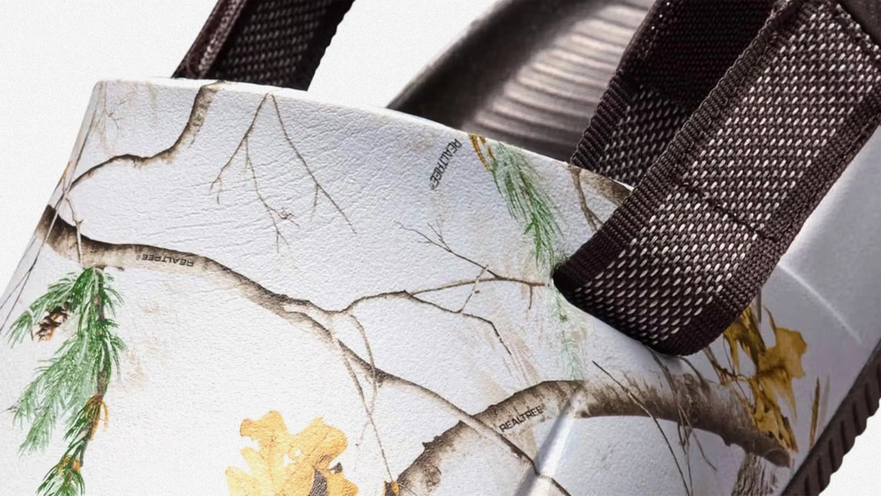 Close-up of a shoe with a white base featuring a camouflage pattern of tree branches, green pine needles, and yellow leaves. The shoe has dark brown woven straps attached to the upper part. The word "REALTREE" is visible within the camouflage design.