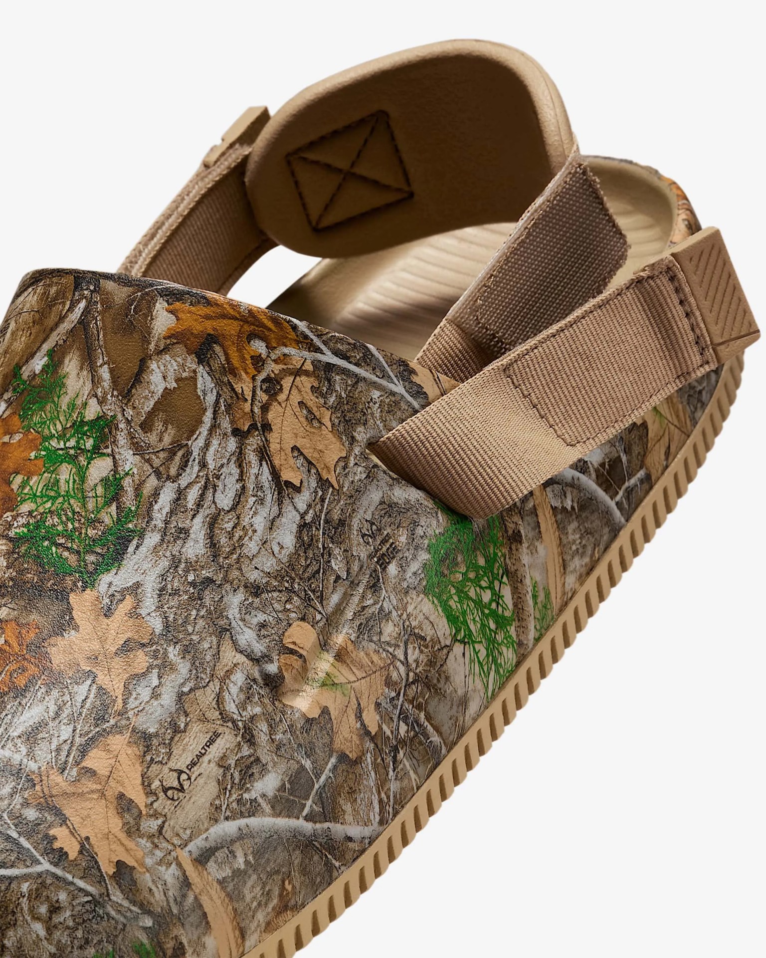 Close-up of a slip-on clog with a camouflage pattern featuring brown, tan, green, and gray leaves and branches. The shoe has a tan adjustable strap with Velcro at the back and a textured tan sole. The interior is beige with a cushioned footbed.