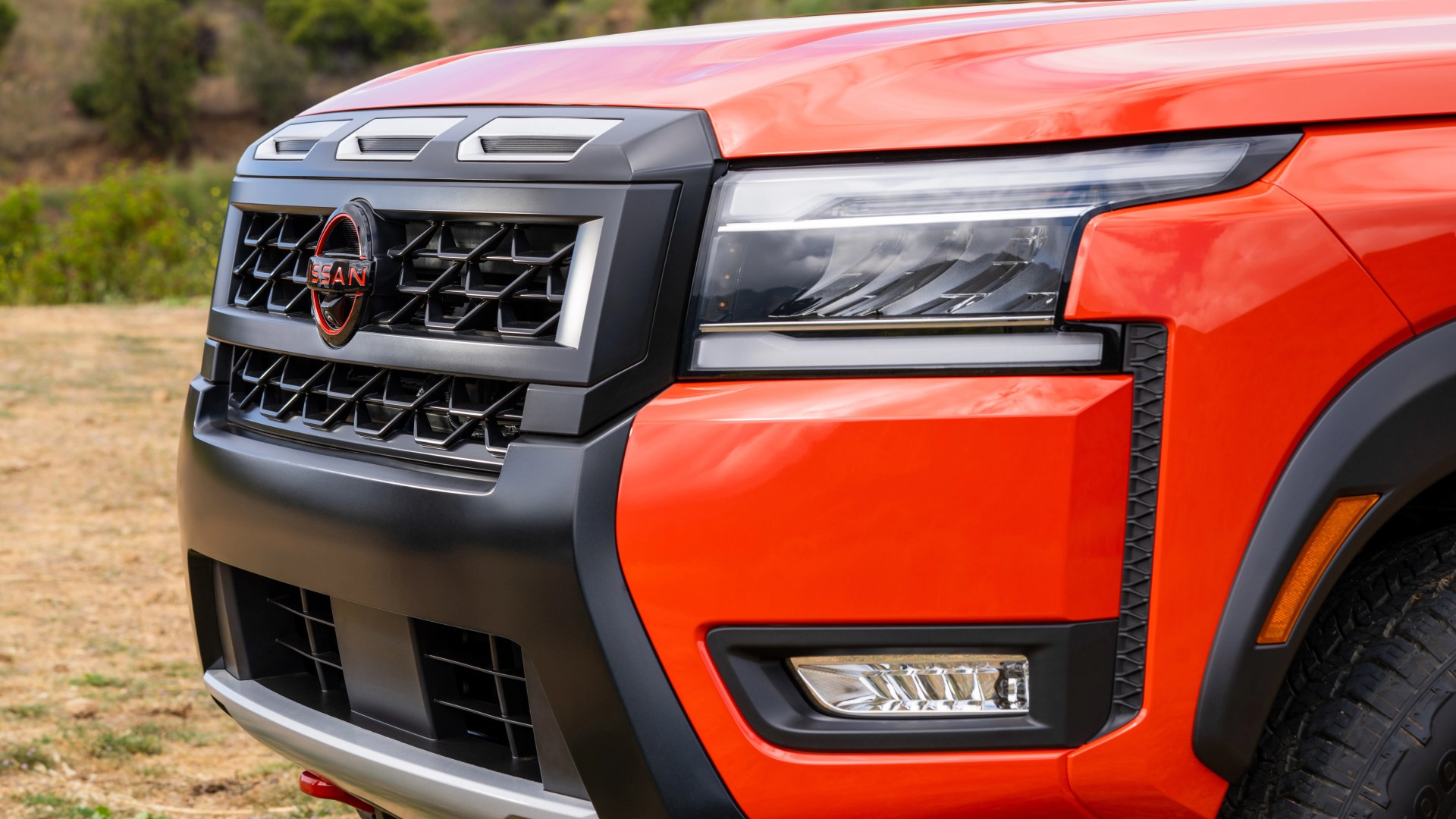 Front right side of an orange Nissan vehicle showing the grille, headlight, and part of the front bumper with black and silver accents. The Nissan emblem is centered on the grille.