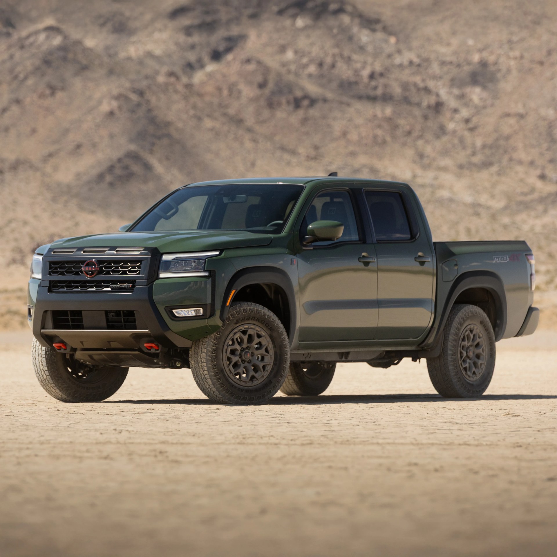 Green Nissan pickup truck with black grille, rugged tires, and red tow hooks, parked on a dry desert landscape with rocky hills in the background.