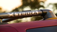 Close-up of a silver roof rack bar on a red vehicle with the word "XTERRA" printed in bold black letters on the bar. The background is blurred with warm, natural lighting.