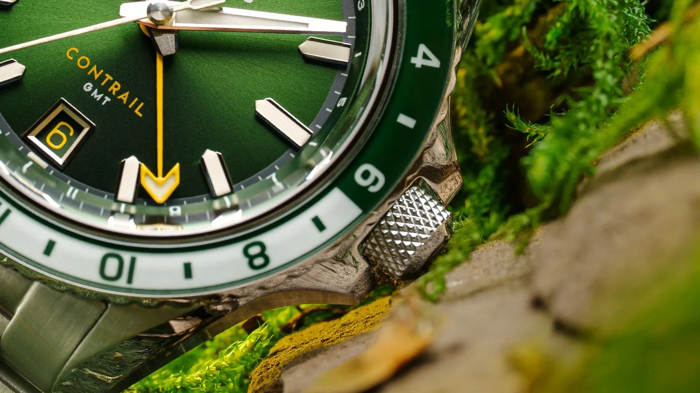 Close-up of a green Nodus Contrail GMT watch face showing the date "6" in a yellow box, with silver hour markers and hands, a yellow GMT hand with an arrow tip, and a green and white bezel marked with numbers. The watch is resting on a surface with green moss and rocks.