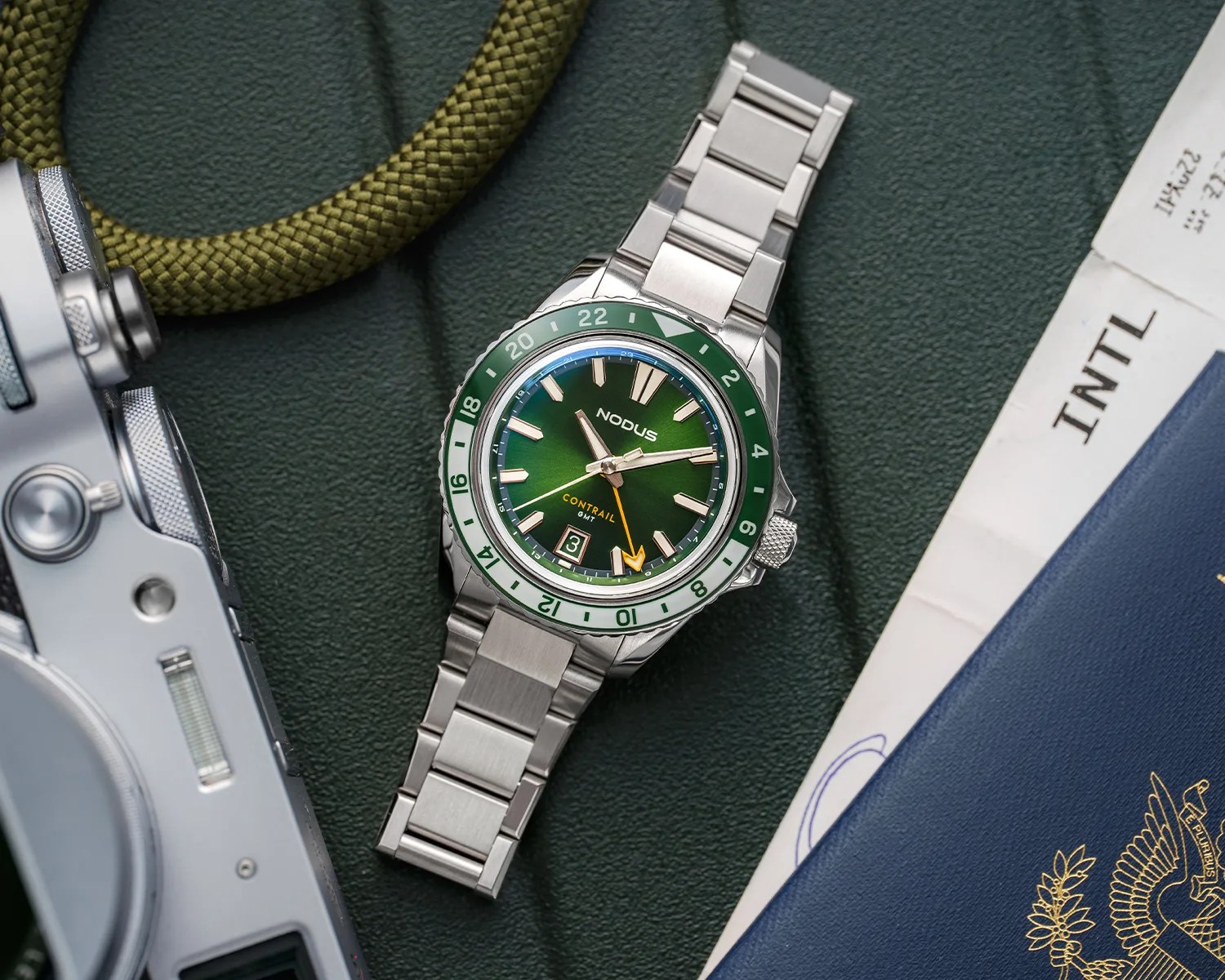 Stainless steel wristwatch with a green gradient dial and matching green and white 24-hour bezel. The watch has silver hour markers and hands, with a yellow GMT hand and a date window at 6 o'clock. The brand name "Nodus" and model "Contrail GMT" are visible on the dial. The watch is placed on a textured dark green surface next to a camera and some documents, including a blue passport.