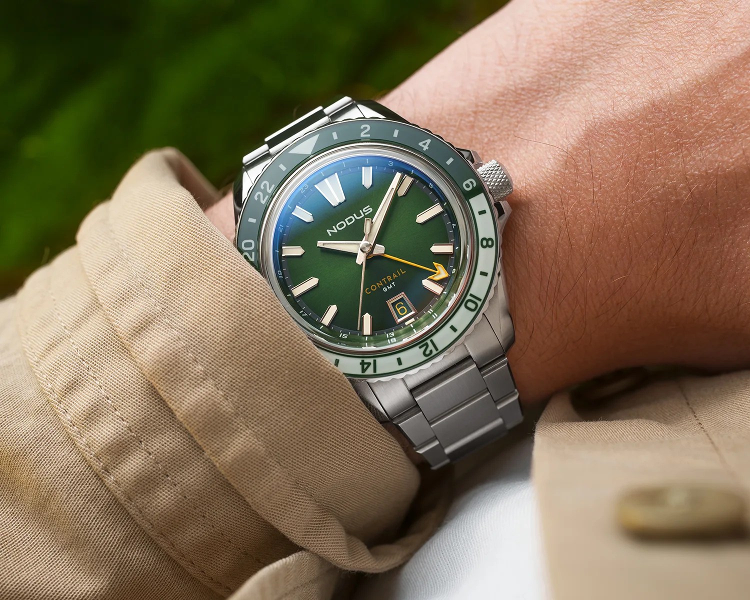 A wristwatch with a green dial and a two-tone green and white bezel, featuring silver hour markers and hands, a yellow GMT hand, and a date window at the 6 o'clock position. The watch has a stainless steel bracelet and the brand name "NODUS" is visible on the dial. The wearer is dressed in a beige jacket.