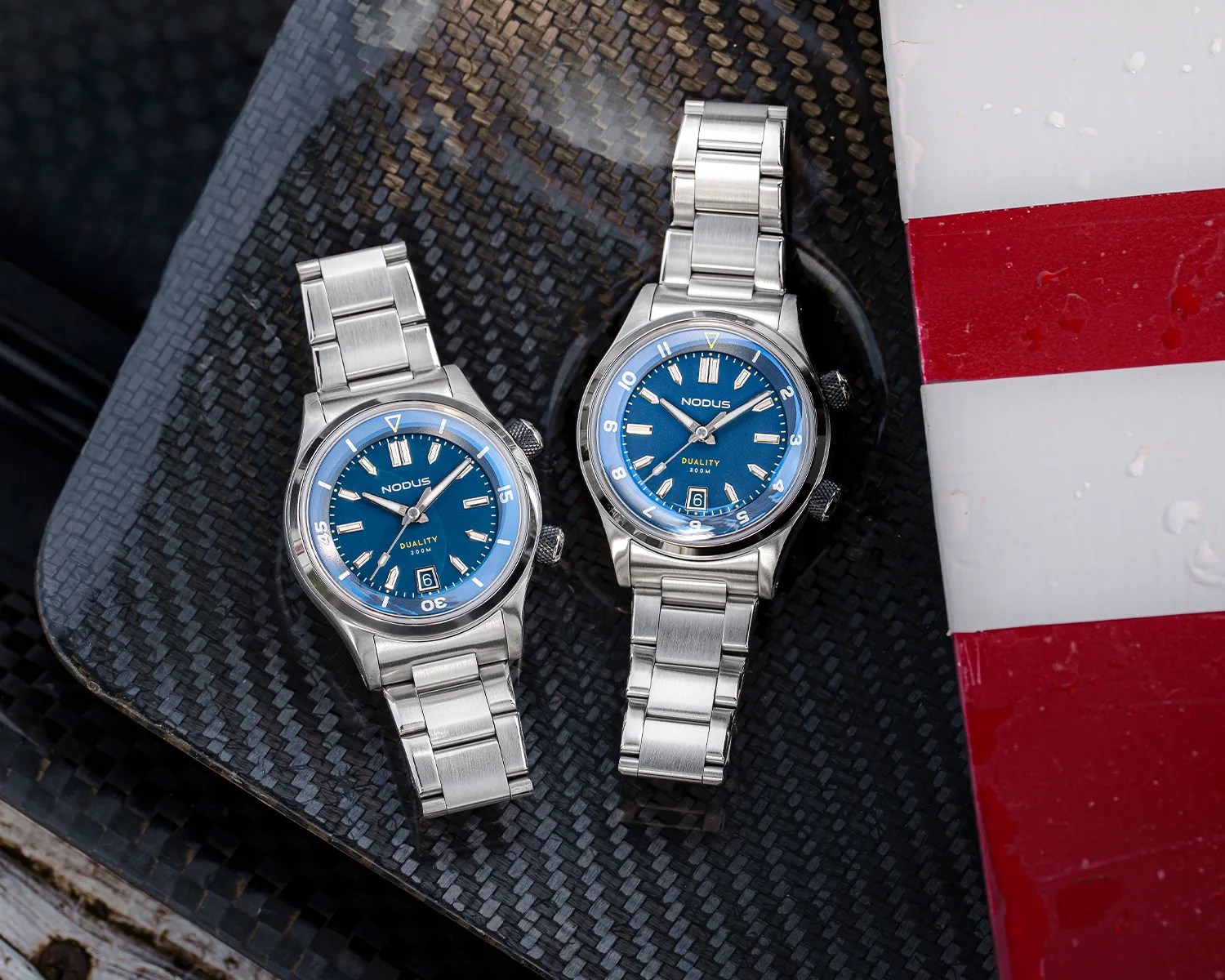 Two stainless steel wristwatches with blue dials and metal link bracelets are placed on a textured black surface. Both watches have the brand name "NODUS" and the model name "DUALITY 300M" on the dial. The watch on the left features a rotating bezel with minute markers, while the watch on the right has a fixed bezel with hour numerals. Both watches display the date at the 6 o'clock position. A red and white striped object is partially visible on the right side of the image.