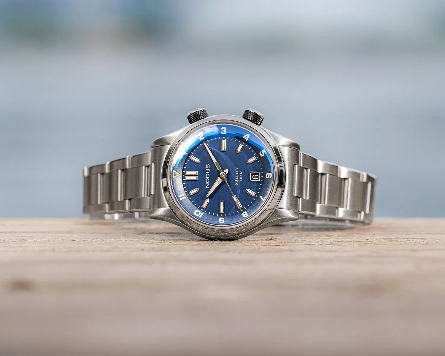 Stainless steel wristwatch with a blue dial, featuring silver hour markers and hands, a date window at the 3 o'clock position, and two black textured crowns. The watch has a metal link bracelet and is labeled "Nodus" and "Duality 300M" on the dial.