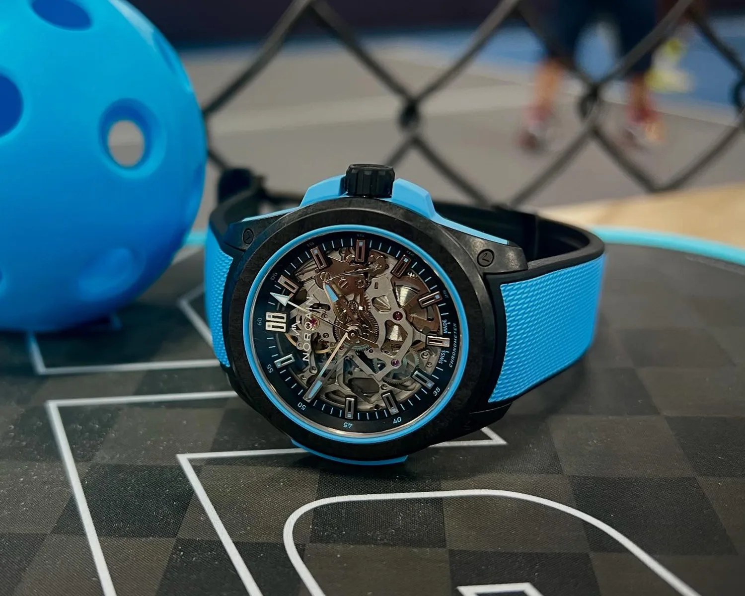A Norqain wristwatch with a blue textured strap and a black bezel. The watch face is skeleton-style, showing the intricate inner mechanical gears and components. The watch is placed on a surface with a black and white geometric pattern, and a blue ball with holes is visible in the background.