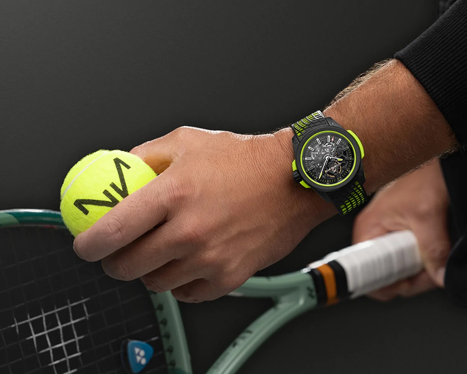 A close-up of a person's hand holding a yellow tennis ball and a tennis racket. The person is wearing a black and neon green Norqain wristwatch with a detailed, partially transparent dial showing the watch's inner mechanics. The tennis racket has a light green frame and a white grip with black and orange accents. The background is dark.