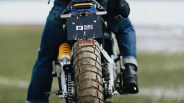 Rear view of a motorcycle with a muddy tire, showing the license plate with the text "NUEN MOTO." The rider is wearing blue jeans and black boots, and the motorcycle has a visible yellow suspension spring. The background is blurred, suggesting an outdoor setting.