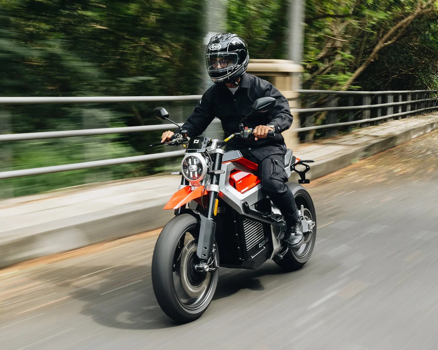 A person wearing a black helmet, black jacket, and black pants is riding a modern electric motorcycle with a red and silver frame on a road with a metal railing and greenery in the background.