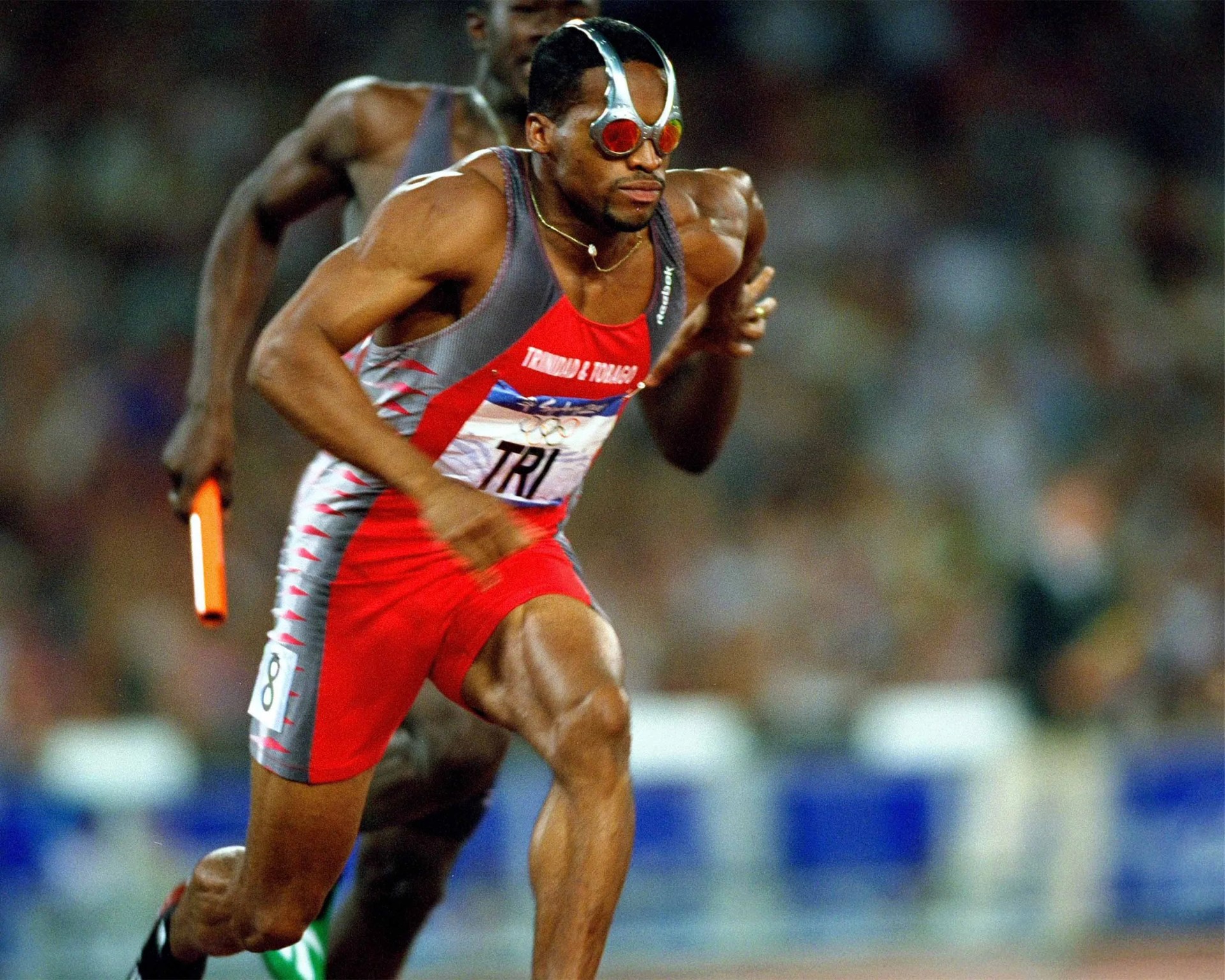 A male sprinter wearing a red and gray Trinidad & Tobago uniform with the number 8 on his shorts is running in a relay race, holding a baton. He is wearing distinctive red-tinted goggles with a silver frame and appears focused and muscular. Another runner is visible behind him, slightly out of focus. The background shows a blurred crowd in a stadium.