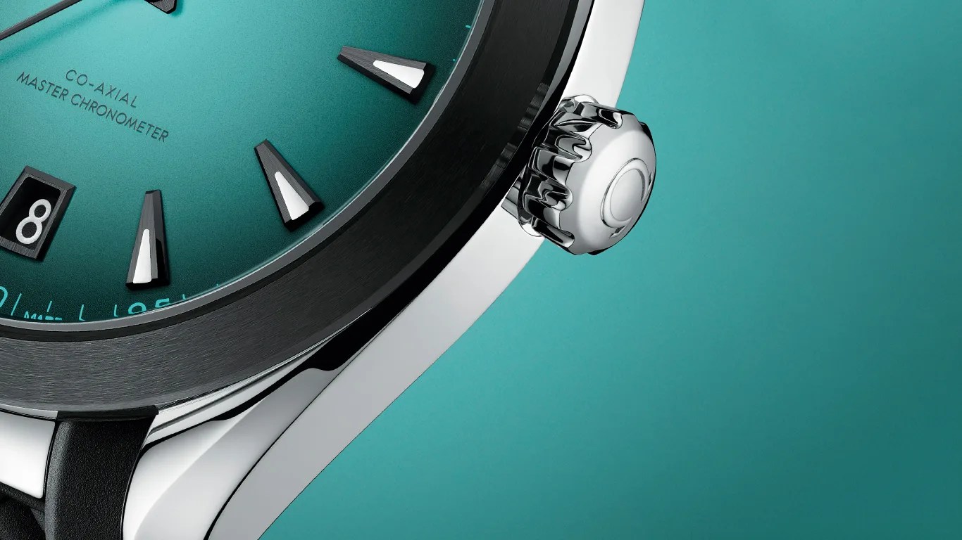 Close-up of an omega watch face with a teal dial, black and white hour markers, and a date window showing the number 8. The watch has a polished silver crown and a black bezel. The text "CO-AXIAL MASTER CHRONOMETER" is visible on the dial.